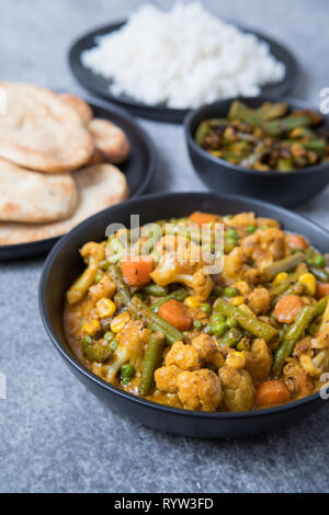 Indian vegetable korma curry in a black bowl in the foreground with Indian side dishes in the background. White rice, naans and okra in a light grey b Stock Photo