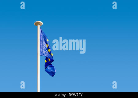 Flag of the European Union (EU) dangling at full mast on a white pole against blue sky. Stock Photo