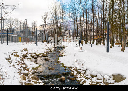Nature park in the city covered with snow through which a mountain stream flows, on the shore there are stones. Visible people walking and benches. Th Stock Photo