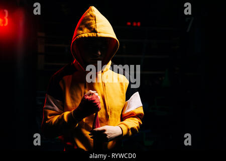 Woman boxer in hood pulls red bandages on hands. Stock Photo