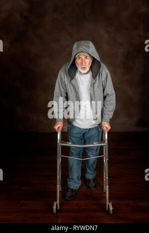 Vertical shot of elderly man resting for a minute while walking with the aid of a walker across a wooden floor.  Brown background.  Hood on his jacket Stock Photo