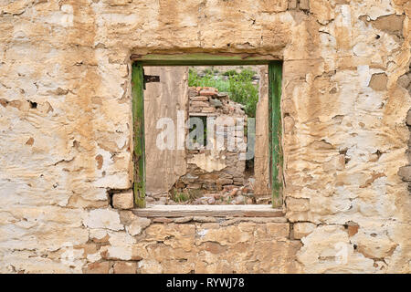 Central empty green window frame on rocky wall of abandoned house.  Through frame see crumbled walls of remaining ruined house Stock Photo