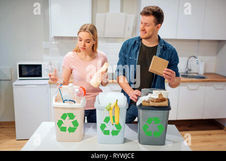 Waste sorting at home. Smiling young family putting plastic, paper, other waste in garbage bio bins in the kitchen Stock Photo