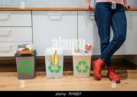 Waste sorting. Cropped view of colorful garbage bins filled with plastic, bio food, paper near womans legs in the kitchen Stock Photo