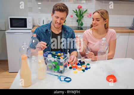 Protect the environment. Aware young couple recycling empty plastic bottles and lids while sitting at the table with other waste at home Stock Photo