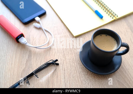 The smartphone is charging from the poverbank. Cup of coffee, diary, pencil and glasses on the table close-up Stock Photo