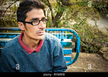 Outdoor image of Asian, Indian serene bespectacled young adult man sitting on park bench and thinking with blank expression while looking away in day Stock Photo