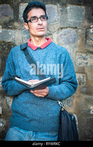 Asian, Indian serene young adult businessman holding business planner and thinking while looking away at view. He is standing with the support of wall Stock Photo