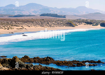 Blue ocean below with a close by rocky shore, white waves rolling on sandy beach beyond under hazy sky. Stock Photo