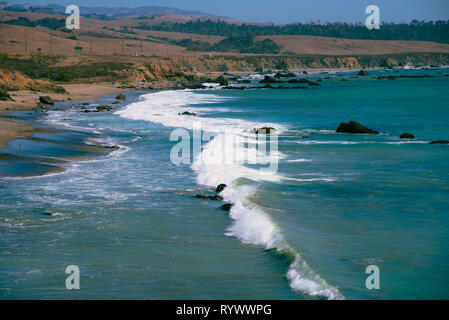 Ocean waves breaking onto shoreline with cliffs and rolling hills beyond under bright blue skies. Stock Photo