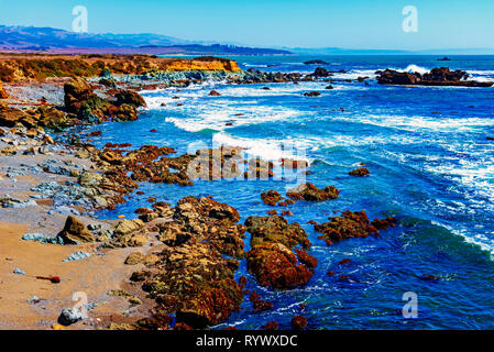 Bright blue ocean,  waves breaking onto rocky shoreline with cliffs under bright blue sky. Stock Photo