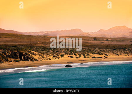 Hazy yellow orange afternoon sky over hazy mountains and golden sandy beach with blue ocean. Waves crashing onto the beach.