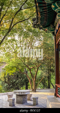 Late afternoon sun shines on a stone table and stools in front of a Chinese temple with trees in the background Stock Photo