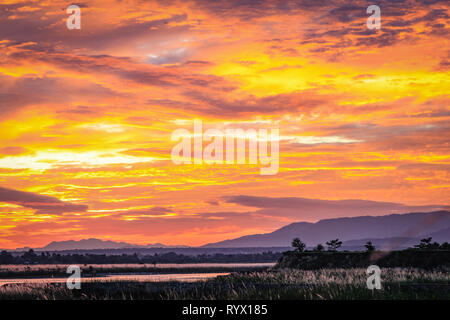 Bright orange and yellow sunset over a flat river with a plateau and small trees on the right Stock Photo
