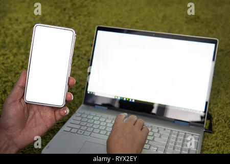 Young man working on laptop and smartphone at home. Modern laptop and cellphone with white blank screens. Freelancer uses mobile and wifi to make busi Stock Photo