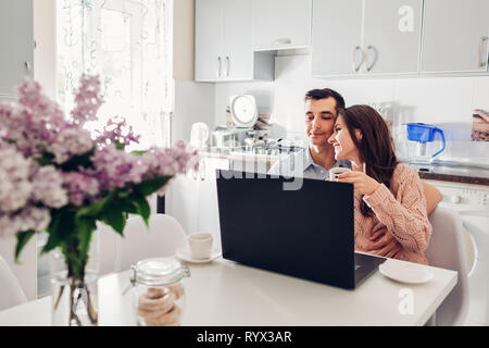 Happy young couple using laptop while having breakfast in modern kitchen. Young man and woman drink coffee and hug. Stock Photo
