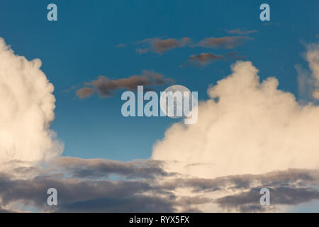 Full moon appears in bright blue sky  framed by white puffy clouds and a layer of dark storm clouds below Stock Photo
