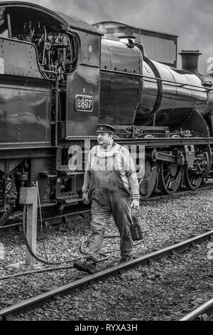 Black & white side view of vintage UK steam locomotive stationary on heritage railway track as train driver, in overalls & cap walks by to change lamp. Stock Photo