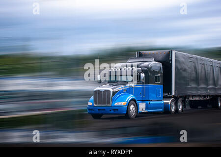 Stylish black and blue big rig long haul semi truck with aerodynamic spoilers transporting commercial cargo in full size black covered semi trailer mo Stock Photo