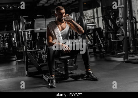 Bodybuilding. Bearded man sitting on bench at gym drinking water relaxed Stock Photo