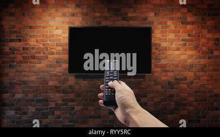 Television on brick wall with hand using remote control Stock Photo