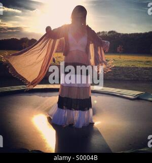 A young woman in a floaty dress bounces on a trampoline at sunset.