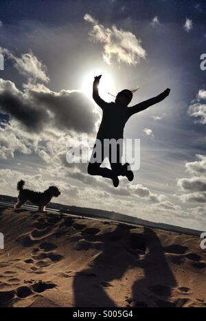 A woman jumps for joy on a sunny day at the beach in Devon, UK. Beautiful blue skies and white fluffy clouds are seen in the background as the lady is silhouetted against the sunshine Stock Photo
