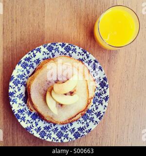 Stack of American pancakes topped with slices of apples on a vintage plate, with a glass of orange juice Stock Photo