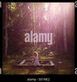 A young girl playing in a sandbox in a dreamlike sun drenched forest Stock Photo