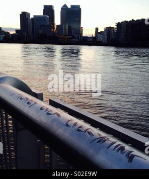 View of Canary Wharf from North Greenwich. Skyscrapers in the background, river in the middle ground and a graffiti in the foreground handrail that reads: 'Money over everything' Stock Photo