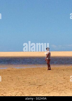 Man  with shorts and teeshirt and  cap on Ocean beach by inland water lake looking out to sea Stock Photo