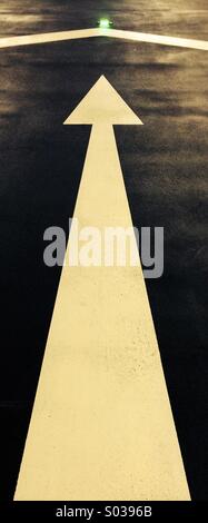 Long white arrow painted on asphalt road pointing at a green cats eye. Stock Photo