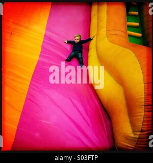 A young two year old boy sliding down aim inflatable slide at a fun fair. The child is showing some degree of fear. Stock Photo