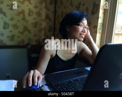 Asian woman smiling and looking out a window while using a laptop computer at home Stock Photo