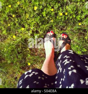 Looking down at feet in sandals and a summer dress standing on a LAN with small yellow flowers