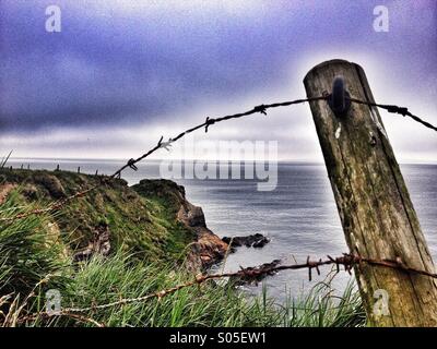 Broken barbed wire fence with cliff top cove in background Stock Photo