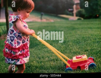 A toddler girl cuts the grass with a toy lawn mower Stock Photo