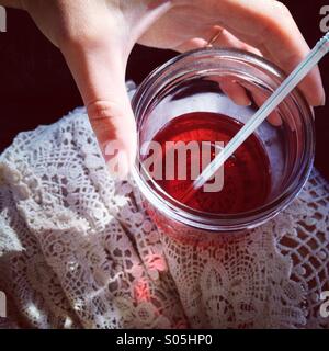 Close up of girl's hand and red drink in glass jar with straw resting on leg in lace dress in summer with sunlight. Stock Photo