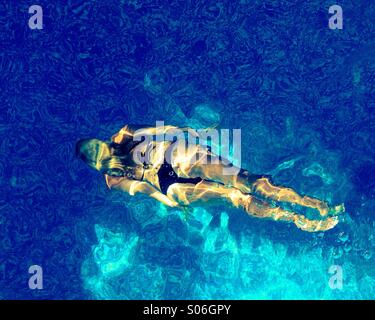 Blond, long haired woman in bikini swimming underwater in the clean warm waters of an outdoor swimming pool illuminated by sunshine. Capture from above. Stock Photo
