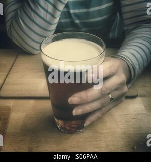 Woman sitting alone at table in public house bar, holding pint glass of beer Stock Photo