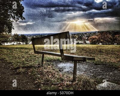 Bench on top of a hill looking out onto suns rays bursting through cloud Stock Photo