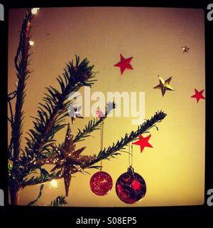 Detail of a Christmas tree decorated with baubles and fairy lights, with red and gold stars in the background Stock Photo