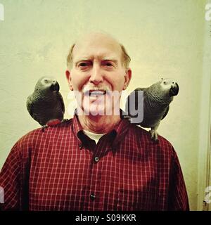 A senior Caucasian man smiles while showing off his two pet African Gray parrots on his shoulders. Stock Photo