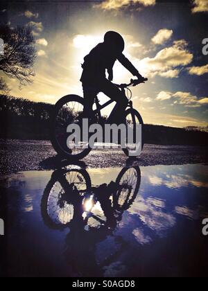 Young girl on bike in silhouette reflected in puddle Stock Photo