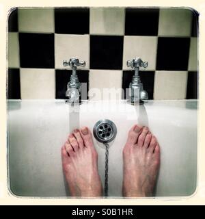A good soak. Pair of man's feet under a pair of hot and cold taps, black and white tile background. Stock Photo