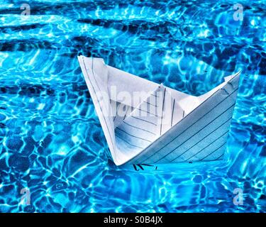 Paper boat folded from a notebook's page, floating on bright blue waters of a swimming pool. Stock Photo