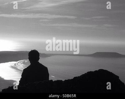 Teenager chilling on rock admiring the view of  the sea surrounding Worms head, Gower. Rear view silhouette