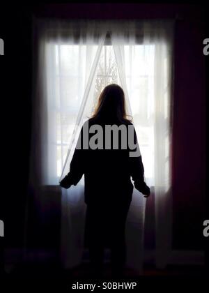 A woman standing at a window, looking out. Stock Photo