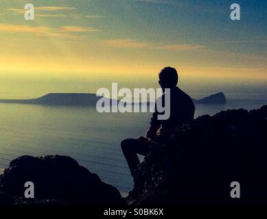 Teenager sitting on a rock overlooking  the sea near Worms head, Gower. Side view silhouette.