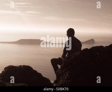 Teenager sitting on a rock overlooking  the sea near Worms head, Gower. Side view silhouette.